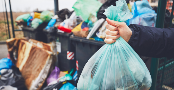 person holding bag of garbage about to throw it into a dumpster overfilling with trash